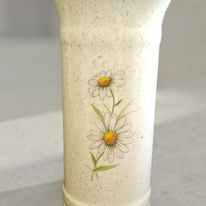 May include: A cylindrical ceramic vase with a cream-colored speckled finish and a gold-trimmed rim. The front of the vase features a botanical illustration of two white daisy flowers with yellow centers and green stems and leaves.