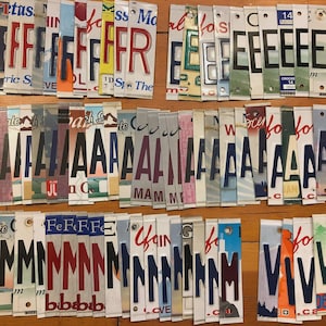 May include: A selection of cut-out letters and numbers from license plates, displayed on a wooden surface. The letters are in diverse colours and fonts, some with state names visible. The arrangement is organised in rows.