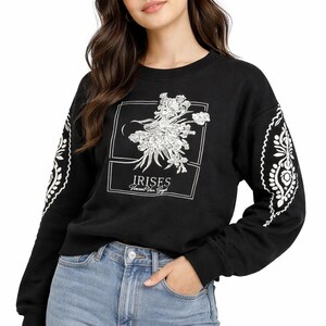 Puede incluir: Sudadera negra con un diseño floral y el texto "IRISES Vincent Van Gogh". Las mangas tienen un estampado floral blanco. La persona lleva vaqueros azules.