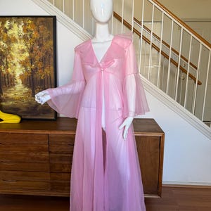May include: A sheer pink vintage robe with bell sleeves and a ruffled collar is displayed on a mannequin. The robe is open in the front and has a flowing, lightweight design. The mannequin is standing in front of a wooden dresser and a staircase.
