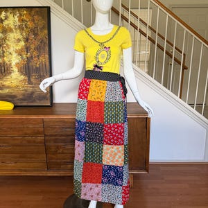 May include: A yellow short-sleeved t-shirt featuring a printed graphic of a necklace and a long wrap skirt with a multi-colored patchwork floral pattern and a black and white polka dot waistband, displayed on a white mannequin.