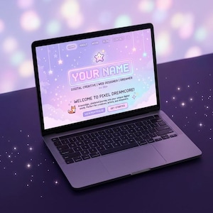 May include: A silver laptop displaying a website design with a pastel purple and blue color scheme. The website features the text "YOUR NAME" and "WELCOME TO PIXEL DREAMCORE!" along with a fox graphic. The laptop is open on a dark surface.