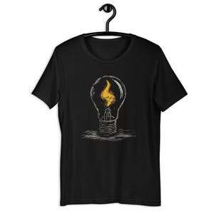 Op de afbeelding: Een zwart t-shirt met korte mouwen en een grafische print van een gloeilamp met een gele vlam erin. Het ontwerp is uitgevoerd in een schetsmatige witte lijnstijl, die het gloeidraad en de glasstructuur van de lamp duidelijk weergeeft.