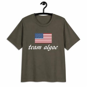 May include: A dark olive green short-sleeve t-shirt featuring a graphic of the United States flag centered on the chest. Below the flag, the text "team algae" is printed in a white, italicized, serif font. The shirt has a crew neckline.