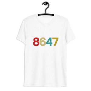May include: White short-sleeved t-shirt featuring the number sequence 8647 across the front. The numbers are in red, gold, teal, and mustard yellow. The t-shirt is hanging on a black hanger.