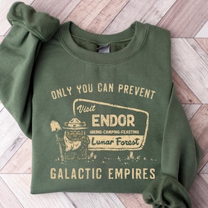 May include: Dark green sweatshirt featuring the text "ONLY YOU CAN PREVENT Visit ENDOR Hiking-Camping-Feasting Lunar Forest GALACTIC EMPIRES". The design includes a bear in a ranger hat. The garment is displayed on a wooden surface with a chevron pattern.