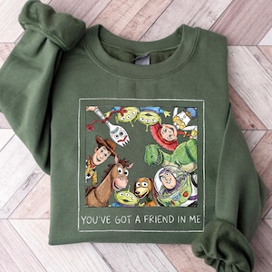 Pu&ograve; includere: Felpa verde oliva con una grafica incorniciata dei personaggi di Toy Story, tra cui Woody e Buzz Lightyear. Il testo "YOU'VE GOT A FRIEND IN ME" &egrave; stampato in bianco sotto. La felpa ha i polsini.