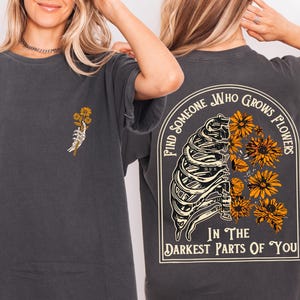 May include: Dark gray t-shirt with a skeleton hand holding yellow flowers on the front. The back of the shirt features a rib cage design with yellow flowers and the text "Find someone who grows flowers in the darkest parts of you."