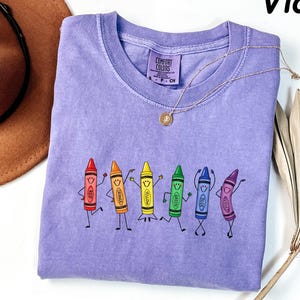 May include: A violet short-sleeved t-shirt featuring a graphic of six anthropomorphic crayons in red, orange, yellow, green, blue, and violet, each with a smiling face and limbs. The shirt has a Comfort Colors label at the inner neckline.