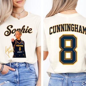 May include: A cream-colored t-shirt featuring a graphic on the front with the name Sophie in script, a basketball player in a navy and gold jersey with the number 8, and a signature. The back displays the name CUNNINGHAM and the number 8.