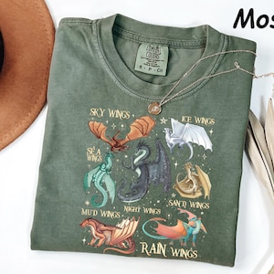 May include: A moss green t-shirt with a graphic of dragon wings, including Sky, Sea, Ice, Mud, Night, Sand, and Rain Wings. The shirt has a crew neck and short sleeves. A gold necklace with a pendant is draped across the shirt.