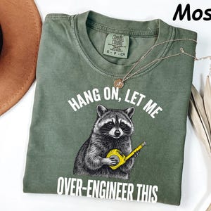 Peut inclure: Un t-shirt à col rond vert mousse avec un motif de raton laveur tenant un mètre ruban jaune. Le texte au-dessus du raton laveur indique HANG ON, LET ME et le texte en dessous indique OVER-ENGINEER THIS. Le t-shirt porte une étiquette Comfort Colors.