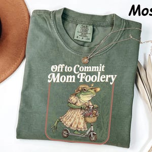 May include: Moss green t-shirt featuring the text "Off to Commit Mom Foolery" and a graphic of a frog on a scooter. The frog is dressed in a dress and hat, and carries a basket. A gold necklace is draped across the shirt.