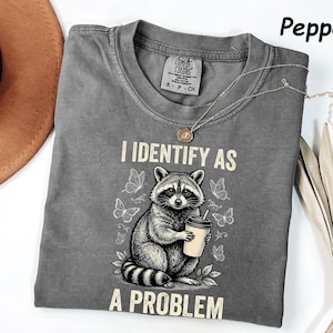 May include: A pepper-coloured t-shirt featuring a graphic of a raccoon holding a coffee cup surrounded by butterflies. The text reads I IDENTIFY AS A PROBLEM. The shirt has a crew neckline and a Comfort Colors brand label visible at the inner collar.