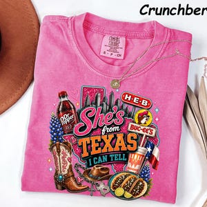 May include: A pink short-sleeved t-shirt featuring a graphic print with the text She's from TEXAS I CAN TELL. The design includes a Dr Pepper bottle, a cowboy boot, a hat, tacos, a Texas map, and logos for H-E-B, Buc-ee's, and Whataburger.