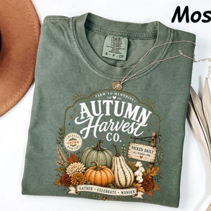 May include: A moss green short-sleeve t-shirt featuring a graphic print with the text Autumn Harvest Co. Farm to Memories. The design includes illustrations of pumpkins, gourds, and autumn flowers, along with a sign reading Picked Daily.