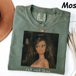 May include: A moss green crew neck t-shirt featuring a graphic print of a doll with long brown hair wearing sunglasses and a pink top. The text Y'all need Jesus is printed in white below the graphic. The shirt has a Comfort Colors brand label.