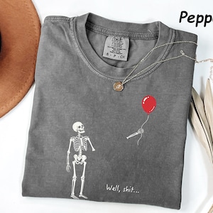 May include: A pepper-colored crew neck t-shirt featuring a white graphic of a skeleton looking at a disembodied skeletal hand holding a red balloon. The text "Well, shit..." is printed in white below the graphic. The shirt has a Comfort Colors label.