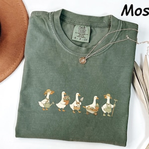 May include: A moss green t-shirt with a crew neck and short sleeves. The shirt features a whimsical illustration of five ducks dressed as gardeners, each with gardening tools. The text "Moss" is in the upper right corner.