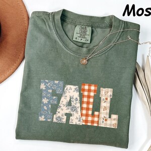 May include: A moss green short-sleeve t-shirt with a crew neckline. The front features the word FALL in large block letters, each with a different pattern: blue floral, cream floral, orange gingham, and a plaid print with small floral accents.