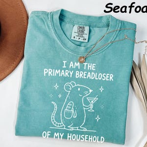 May include: Seafoam green t-shirt with the text "I AM THE PRIMARY BREADLOSER OF MY HOUSEHOLD" and a cartoon mouse holding a cocktail and a loaf of bread. The shirt has a crew neck and short sleeves. A gold necklace is draped across the shirt.