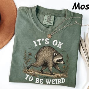 May include: A moss green short-sleeve t-shirt featuring a graphic of a raccoon walking among flowers and mushrooms. The text "IT'S OK TO BE WEIRD" is printed in white above and below the raccoon. The shirt has a Comfort Colors brand label.