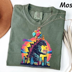 May include: Moss green Comfort Colors t-shirt featuring a colorful graphic of Godzilla holding a birthday cake, with balloons and a city skyline. The shirt has a crew neck and short sleeves. The word "Moss" is in the upper right corner.