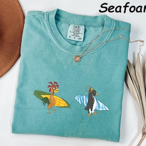 May include: Seafoam green t-shirt with a crew neck and short sleeves. The shirt features a cartoon rooster and penguin, each riding a surfboard. A gold necklace with a pendant is draped across the shirt.