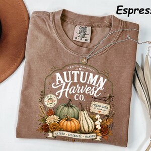 May include: An espresso-colored short-sleeve t-shirt featuring a graphic print with the text "Autumn Harvest Co." and "Farm to Memories." The design includes illustrations of pumpkins, gourds, autumn flowers, and a sign reading "Picked Daily."