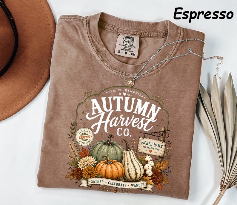 May include: An espresso-colored short-sleeve t-shirt featuring a graphic print with the text "Autumn Harvest Co." and "Farm to Memories." The design includes illustrations of pumpkins, gourds, autumn flowers, and a sign reading "Picked Daily."