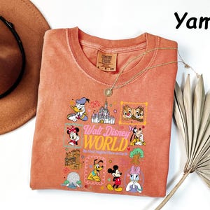 K&ouml;nnte beinhalten: Ein korallenfarbenes T-Shirt mit einer Grafik verschiedener Disney-Figuren und dem Text "Walt Disney World". Das Shirt hat einen Rundhalsausschnitt und kurze &Auml;rmel. Eine goldene Halskette ist &uuml;ber dem Shirt drapiert. Ein brauner Hut befindet sich in der oberen linken Ecke.