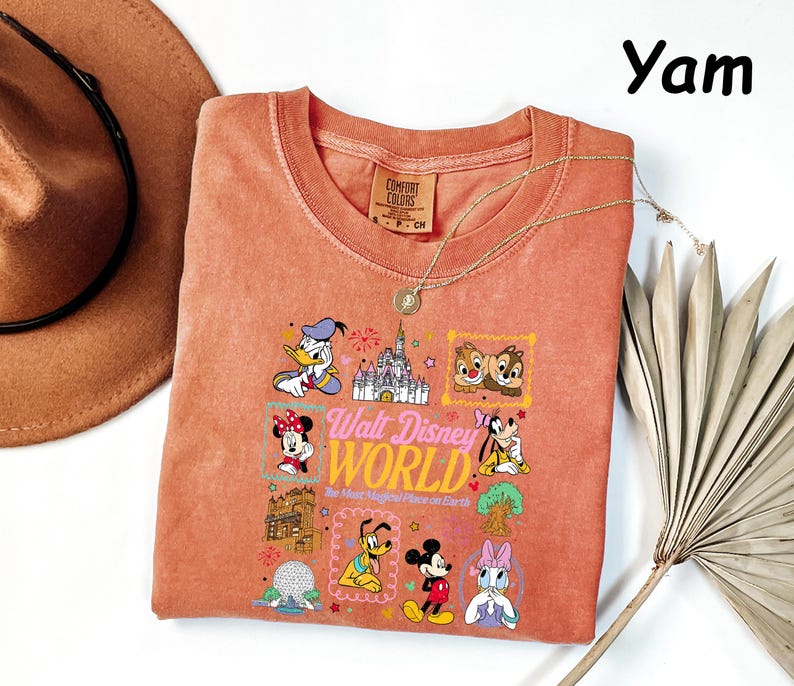 K&ouml;nnte beinhalten: Ein korallenfarbenes T-Shirt mit einer Grafik verschiedener Disney-Figuren und dem Text "Walt Disney World". Das Shirt hat einen Rundhalsausschnitt und kurze &Auml;rmel. Eine goldene Halskette ist &uuml;ber dem Shirt drapiert. Ein brauner Hut befindet sich in der oberen linken Ecke.
