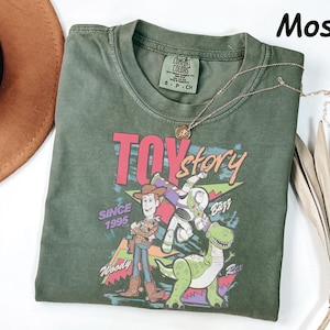 May include: A moss green t-shirt featuring the Toy Story logo and characters Woody, Buzz Lightyear, and Rex. The graphic includes the text "Toy Story" and "Since 1995". A gold necklace is draped across the shirt.