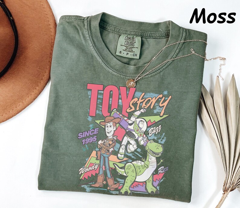 May include: A moss green t-shirt featuring the Toy Story logo and characters Woody, Buzz Lightyear, and Rex. The graphic includes the text "Toy Story" and "Since 1995". A gold necklace is draped across the shirt.