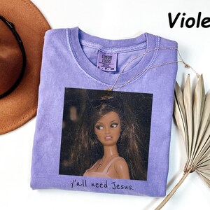 May include: A violet short-sleeved t-shirt featuring a graphic print of a doll with long brown hair and a pink top. The text "Y'all need Jesus." is printed below the graphic. The shirt has a Comfort Colors label at the inner neckline.