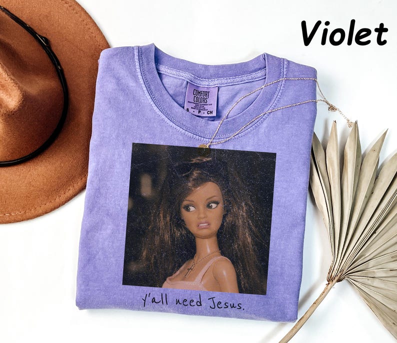 May include: A violet short-sleeved t-shirt featuring a graphic print of a doll with long brown hair and a pink top. The text "Y'all need Jesus." is printed below the graphic. The shirt has a Comfort Colors label at the inner neckline.