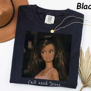 May include: A black t-shirt featuring a graphic print of a doll with long brown hair, wearing sunglasses on her head and a necklace. The text "y'all need Jesus." is printed in white below the graphic. The shirt has a Comfort Colors brand label.