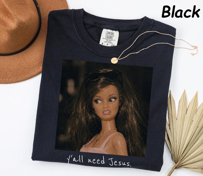 May include: A black t-shirt featuring a graphic print of a doll with long brown hair, wearing sunglasses on her head and a necklace. The text "y'all need Jesus." is printed in white below the graphic. The shirt has a Comfort Colors brand label.