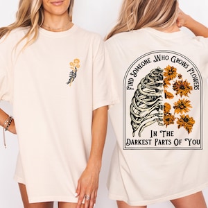 May include: Cream-colored t-shirt with a graphic on the front of a skeleton hand holding yellow flowers. The back of the shirt features a rib cage design with yellow flowers and the text "Find Someone Who Grows Flowers In The Darkest Parts Of You."