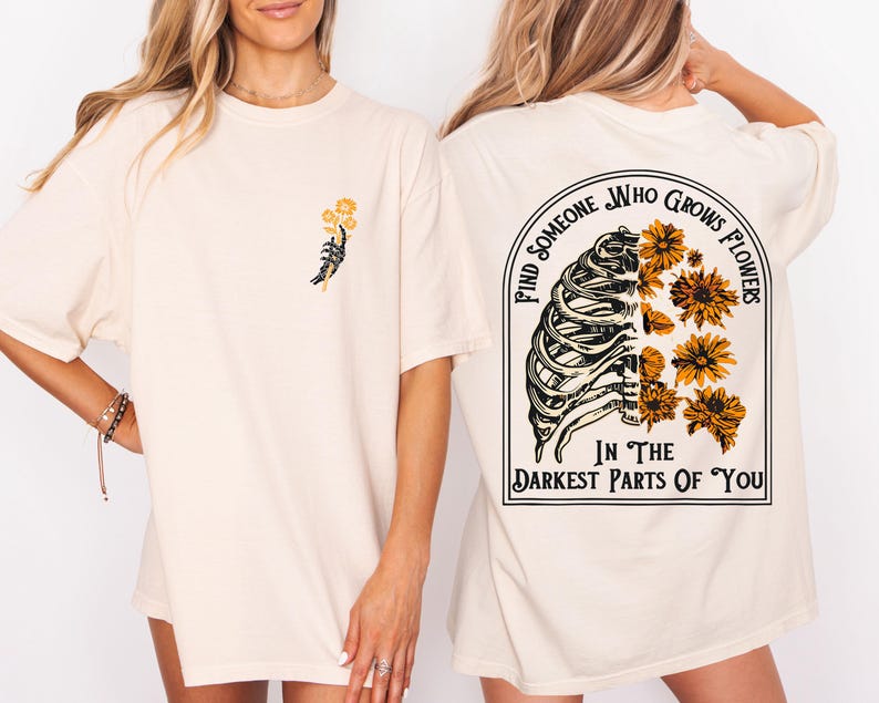 May include: Cream-colored t-shirt with a graphic on the front of a skeleton hand holding yellow flowers. The back of the shirt features a rib cage design with yellow flowers and the text "Find Someone Who Grows Flowers In The Darkest Parts Of You."