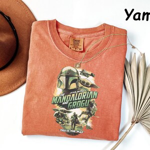 May include: A coral-colored, crew neck t-shirt featuring a graphic from "The Mandalorian and Grogu." The design includes the Mandalorian's helmet, Grogu, and the text "The Mandalorian and Grogu" and "This is the Way."