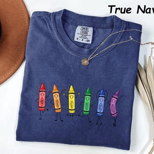 May include: A true navy blue short-sleeve t-shirt featuring a graphic of six anthropomorphic crayons in red, orange, yellow, green, blue, and violet. Each crayon has a smiling face and stick-figure limbs. The shirt has a Comfort Colors label.