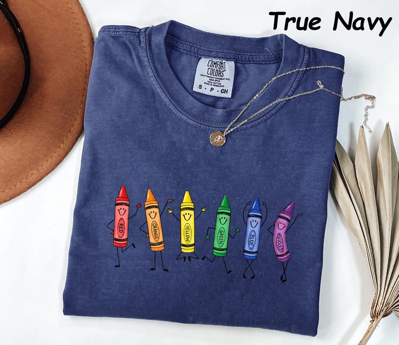 May include: A true navy blue short-sleeve t-shirt featuring a graphic of six anthropomorphic crayons in red, orange, yellow, green, blue, and violet. Each crayon has a smiling face and stick-figure limbs. The shirt has a Comfort Colors label.