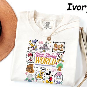 K&ouml;nnte beinhalten: Cremefarbenes T-Shirt mit einer farbenfrohen Grafik von Disney-Figuren und Wahrzeichen. Das Design umfasst Donald Duck, Minnie Mouse, Mickey Mouse, Chip und Chap und den Text "Walt Disney World".