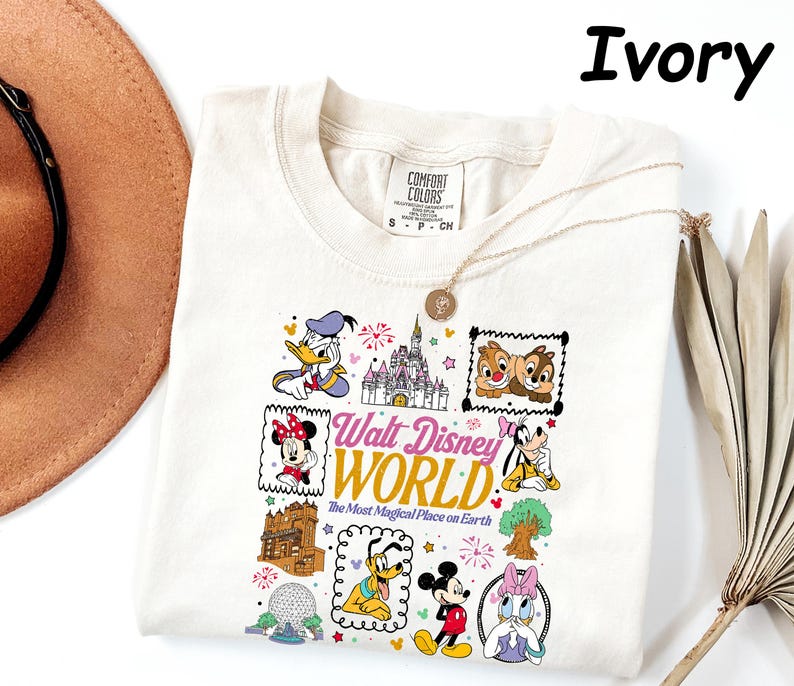 K&ouml;nnte beinhalten: Cremefarbenes T-Shirt mit einer farbenfrohen Grafik von Disney-Figuren und Wahrzeichen. Das Design umfasst Donald Duck, Minnie Mouse, Mickey Mouse, Chip und Chap und den Text "Walt Disney World".