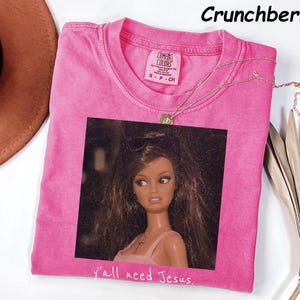 May include: A bright pink short-sleeved t-shirt with a graphic print of a doll with long brown hair and a cross necklace. The text "y'all need Jesus." is printed below the graphic. The shirt has a Comfort Colors label showing size S.