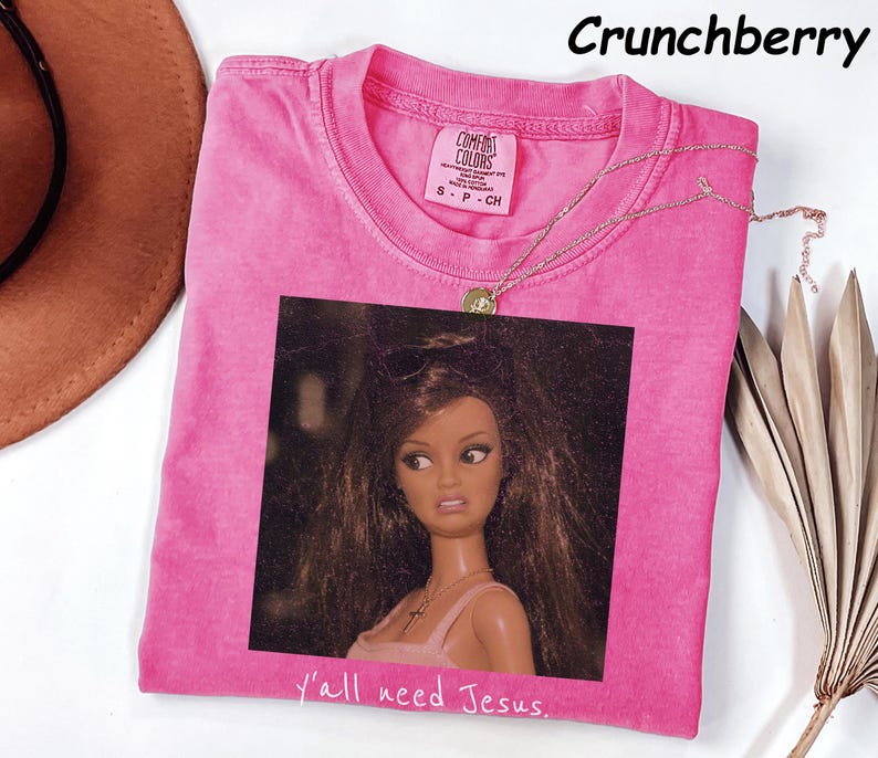 May include: A bright pink short-sleeved t-shirt with a graphic print of a doll with long brown hair and a cross necklace. The text "y'all need Jesus." is printed below the graphic. The shirt has a Comfort Colors label showing size S.