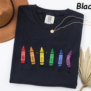 May include: A black crew neck t-shirt featuring a graphic print of six colorful cartoon crayons with faces, arms, and legs. The crayons are labeled red, orange, yellow, green, blue, and violet. The shirt has a Comfort Colors brand tag at the neck.