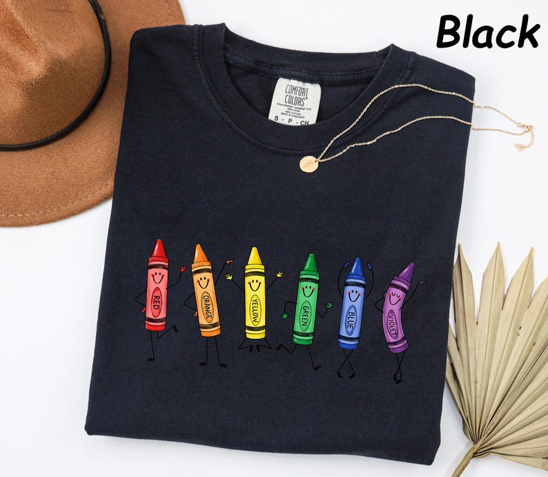 May include: A black crew neck t-shirt featuring a graphic print of six colorful cartoon crayons with faces, arms, and legs. The crayons are labeled red, orange, yellow, green, blue, and violet. The shirt has a Comfort Colors brand tag at the neck.