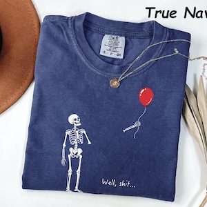 May include: A true navy blue t-shirt featuring a white graphic of a skeleton looking at a detached skeletal hand holding a red balloon. The text "Well, shit..." is printed in white below the graphic. The shirt has a Comfort Colors brand label.
